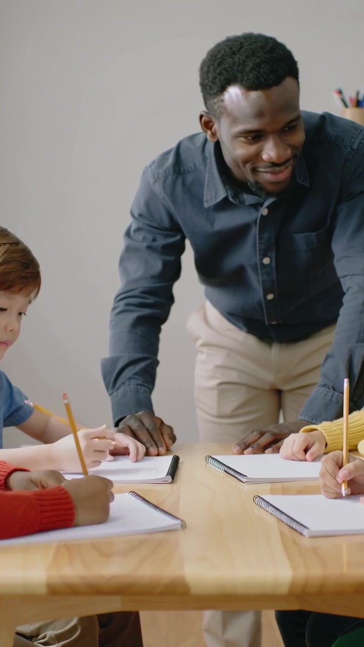 Vertical video: Pointing at page, teacher in blue leaning at school table guiding kids' pencil work