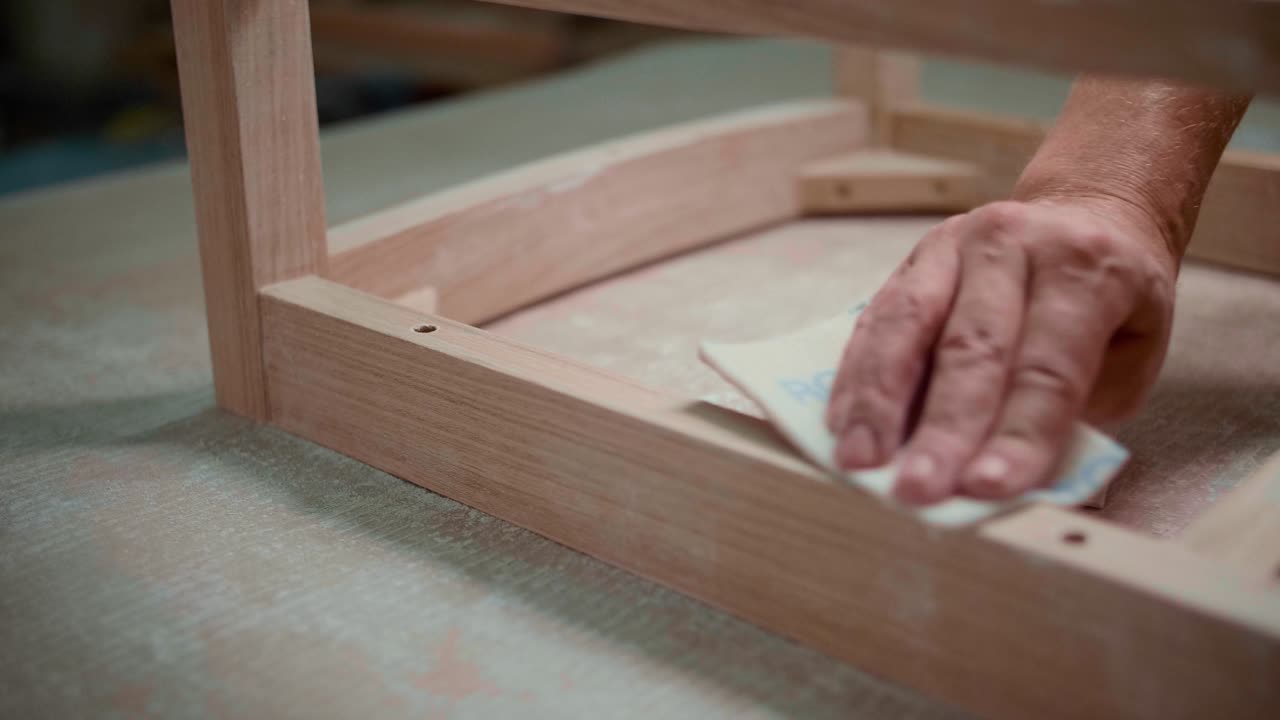 A close-up of a hand sanding a wooden frame with a sanding block, highlighting careful craftsmanship and smooth finishing in woodworking.