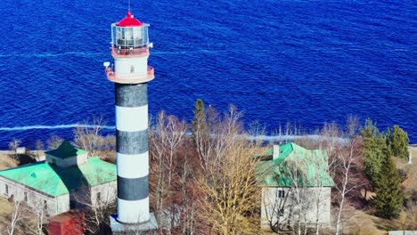 a tall black and white lighthouse stands among simple houses with green roofs along the bright blue baltic sea, framed by leafless trees and clear daylight.
