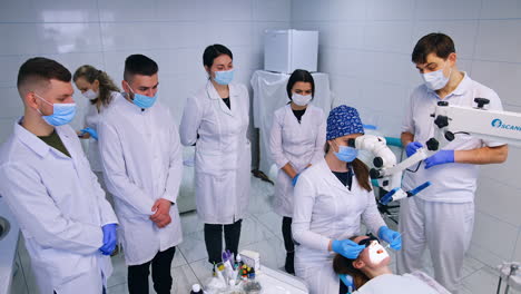 student sits looking at microscope and holding tools in patient's mouth. teacher helps to set the equipment. group of students stand around.