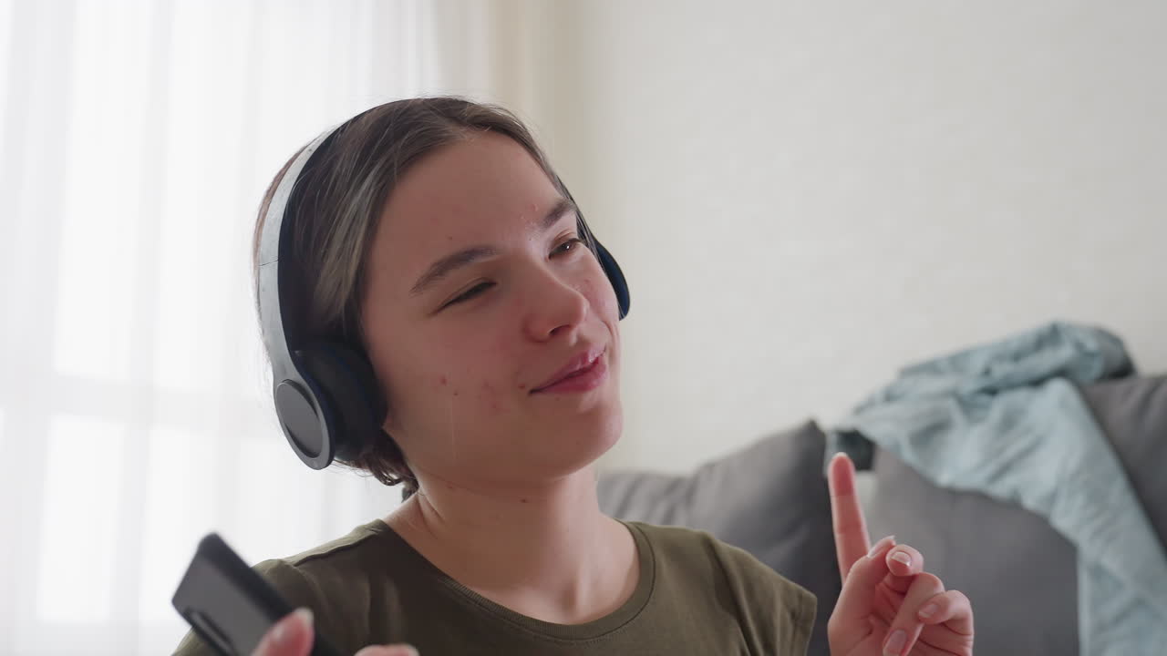 Artista musicale seduto in interni con cuffie e telefono in mano, occhi chiusi, sorridente mentre si muove a ritmo di musica, abbigliamento casual rilassato, divano sullo sfondo con vestiti sparsi