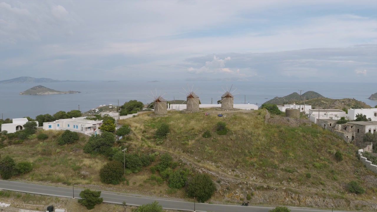 Veduta aerea dei tradizionali mulini a vento di Patmos in cima a una collina, che dominano un villaggio sparso di case bianche e una baia costiera.
