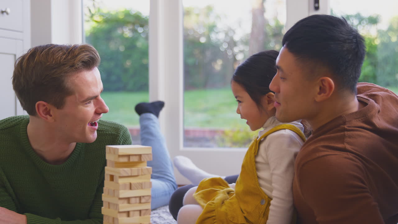 Daughter Whispering In Parent's Ear As Family With Two Dads Play Game Stacking Wooden Bricks