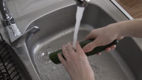 close above shoulder shot as a woman washing a cucumber over a metal sink with fresh water