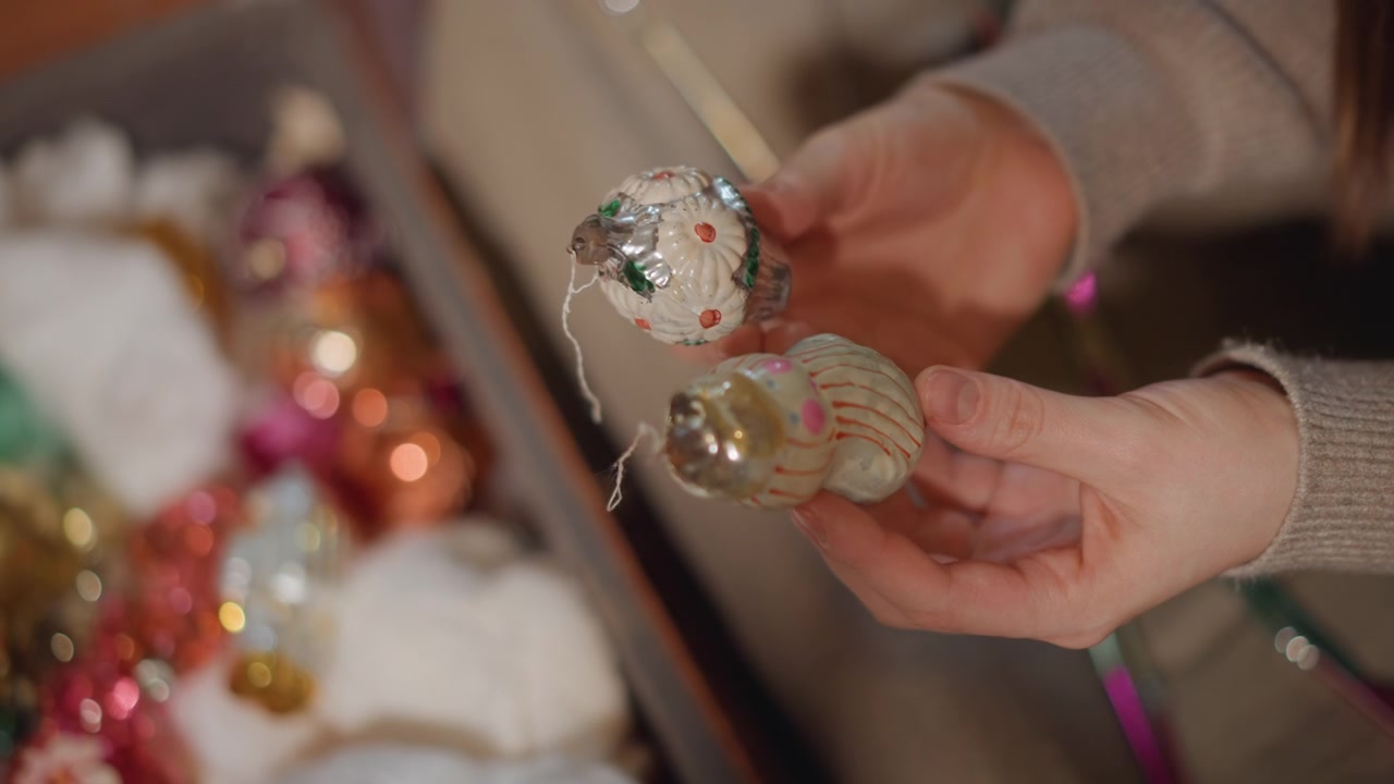 Mani che tengono delicatamente un ornamento di vetro vintage, donna caucasica che ispeziona una fragile pallina in una scatola accogliente del soggiorno con carta velina, luci bokeh calde, dita attente che sistemano lustrini e fili d'argento, tenero.