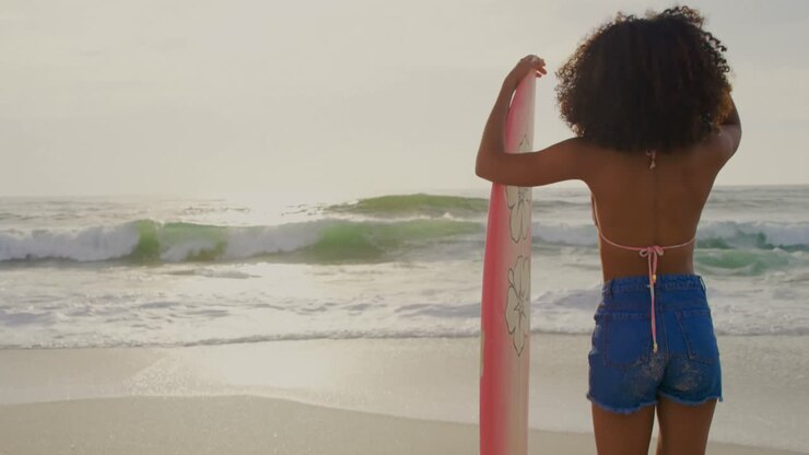 Vista posteriore di surfista afroamericana in piedi con la tavola da surf sulla spiaggia 4K