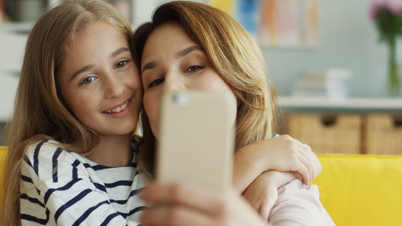 거실 1에 있는 소파에 앉아 스마트폰으로 셀카를 찍는 어머니와 귀여운 소녀의 모습을 가까이에서 봅니다.