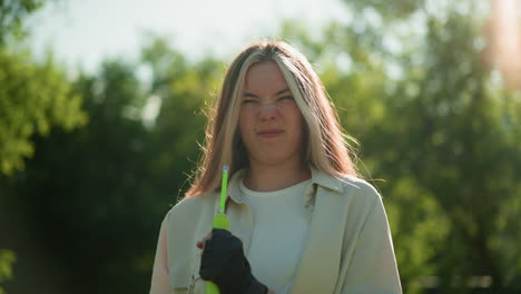 young girl stands with a focused expression, holding a green air pump, shaking it with a gentle breeze blowing her hair against a background of lush trees