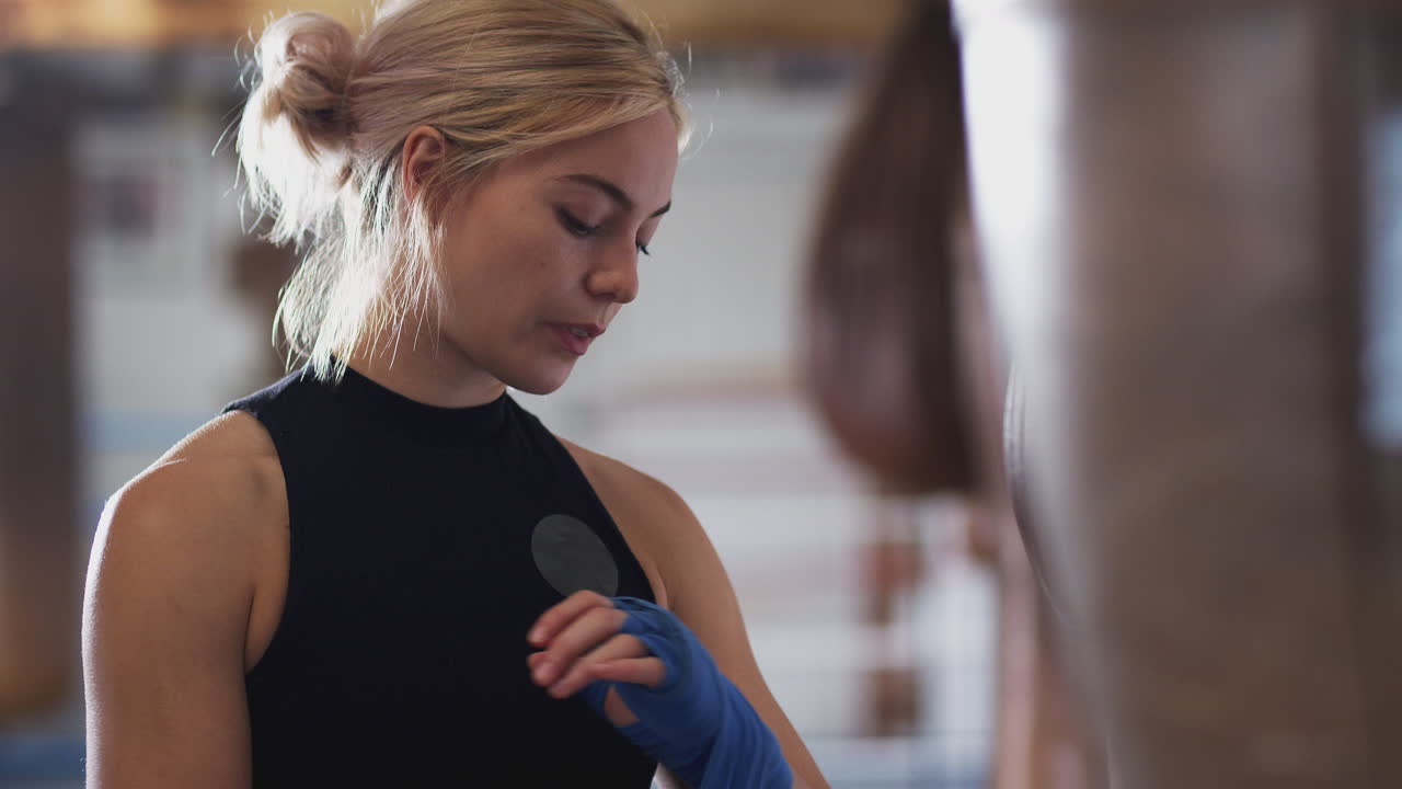 Female Boxer Training In Gym Putting Wraps On Hands Standing Next To Punching Bag