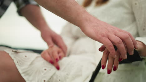 close-up of a man gently positioning a woman's hands on her lap, highlighting her red nails and a unique ring in a tranquil field