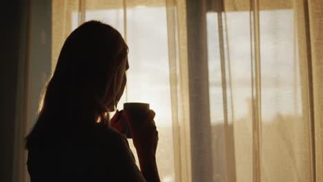 Silhouette-Einer-Frau-Mit-Einer-Tasse-Tee,-Steht-Bei-Sonnenuntergang-Am-Fenster