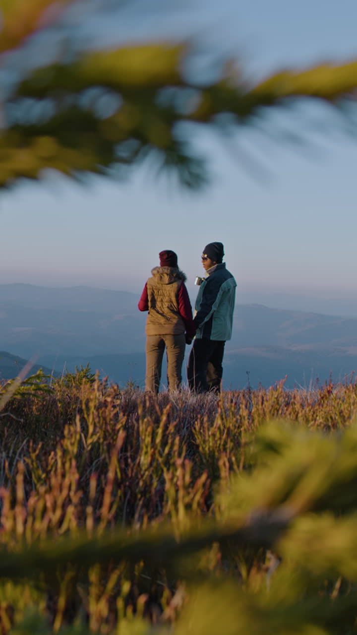 una pareja de excursionistas se toman de la mano, se paran en la cima de una colina y disfrutan de la vista en otoño. un hombre afroamericano con una mujer caucásica beben té y hablan. una joven familia multiétnica en sus vacaciones en las montañas.