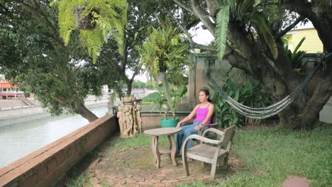 Thailändische-Dame-Sitzt-In-Einem-Hölzernen-Gartenstuhl-Mit-Blick-Auf-Einen-Kanal-In-Ayutthaya,-Thailand