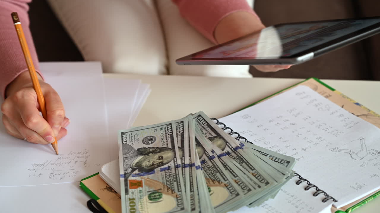 A woman writes calculations in a notebook while holding a tablet, surrounded by sheets of paper and dollar bills