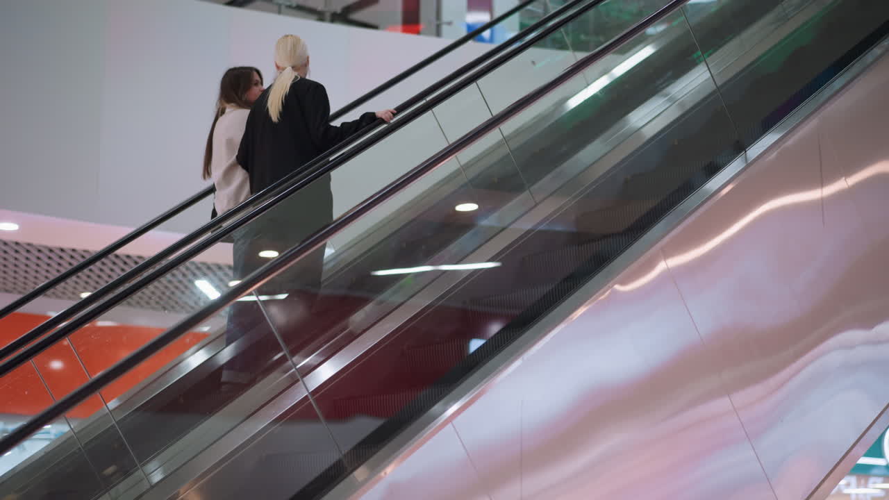 Two women ride ascending escalator inside shopping mall, holding handrail and chatting while going up, surrounded by bright interior lights, modern architecture and retail store signs in background