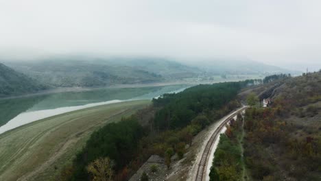 Aerial-view-of-railroad-tracks-along-a-mountain-side,-surrounded-by-trees,-with-river-and-mountains-in-background