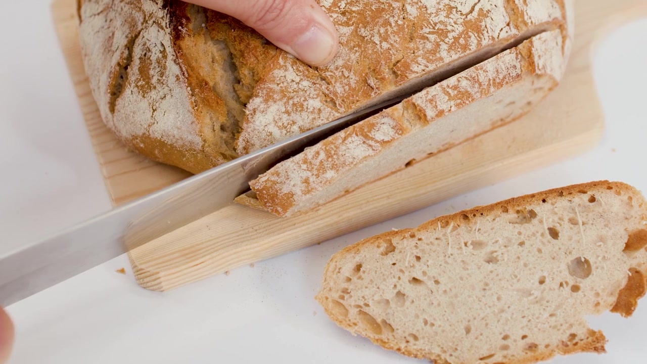 Primo piano di mani che tagliano pane di pasta madre croccante su un tagliere di legno su sfondo bianco