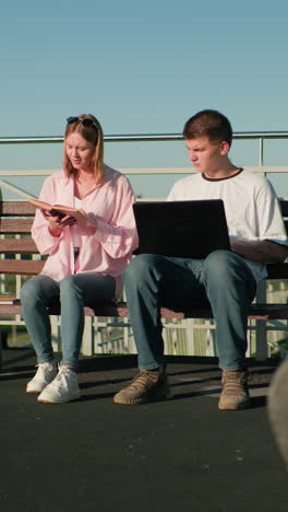 estudiantes masculinos y femeninos estudiando al aire libre en un banco; hombre escribiendo en la computadora portátil mientras se involucra con la pareja femenina leyendo de su libro, en un telón de fondo del horizonte urbano