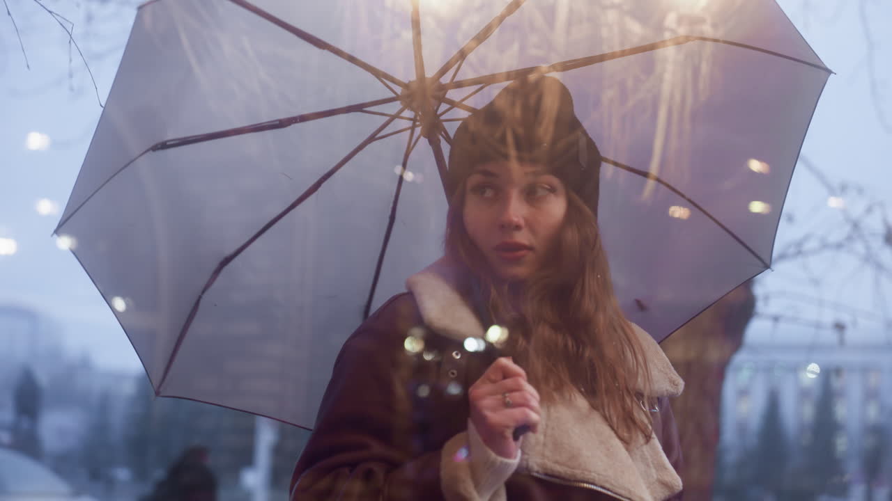 Dreamy girl holding umbrella walking past cafe, looking through glass window on cold overcast day, wearing black knit cap, brown shearling jacket, passing cars, and wet street