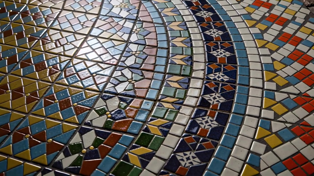 Kamera dras tillbaka och avslöjar en cirkulär keramisk mosaik på ett skyddat golv, som visar glaserade plattor.