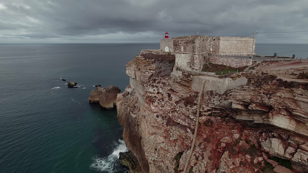 Nazaré, Portekiz'de turkuaz Atlantik suyunun üzerinde deniz feneri ve kaleyi ortaya çıkaran, katmanlı kaya oluşumlarını ve engebeli kıyı şeridini gösteren, Nazaré kayalıkları boyunca yavaş drone dolly kayışı.