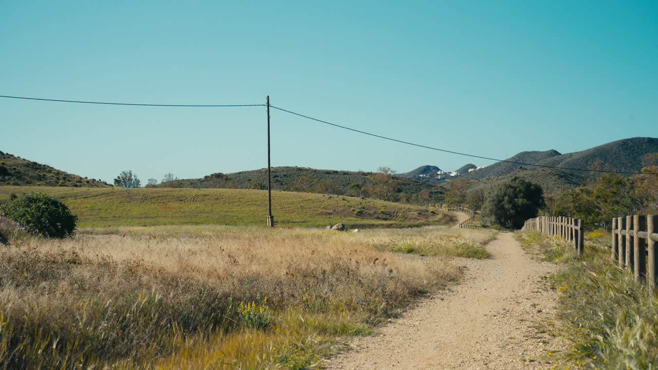 Strada sterrata rurale che attraversa un paesaggio rurale mediterraneo con dolci colline, erba secca e cielo azzurro limpido. Paesaggio di viaggio tranquillo con un senso di viaggio ed esplorazione