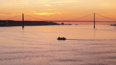 tug boat ferry crosses tagus river at sunset as last light dusk glow spreads on silhouette of suspension bridge