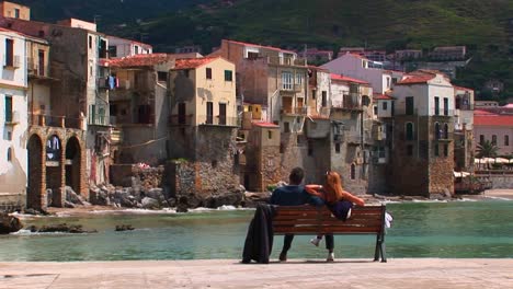 a couple sits on a bench overlooking the ocean and houses in cefalu italy