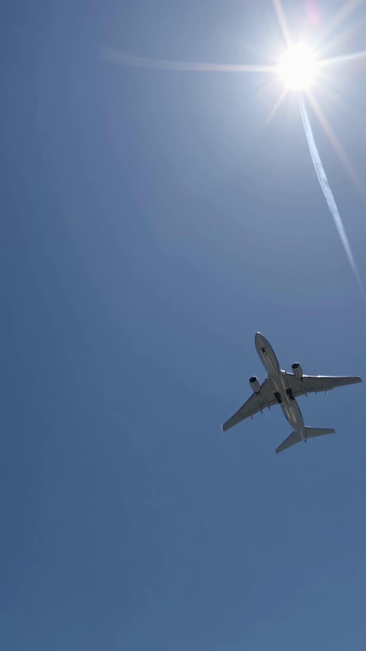 Low-angle video shot of an airplane flying under a bright sun, capturing a contrail against a clear