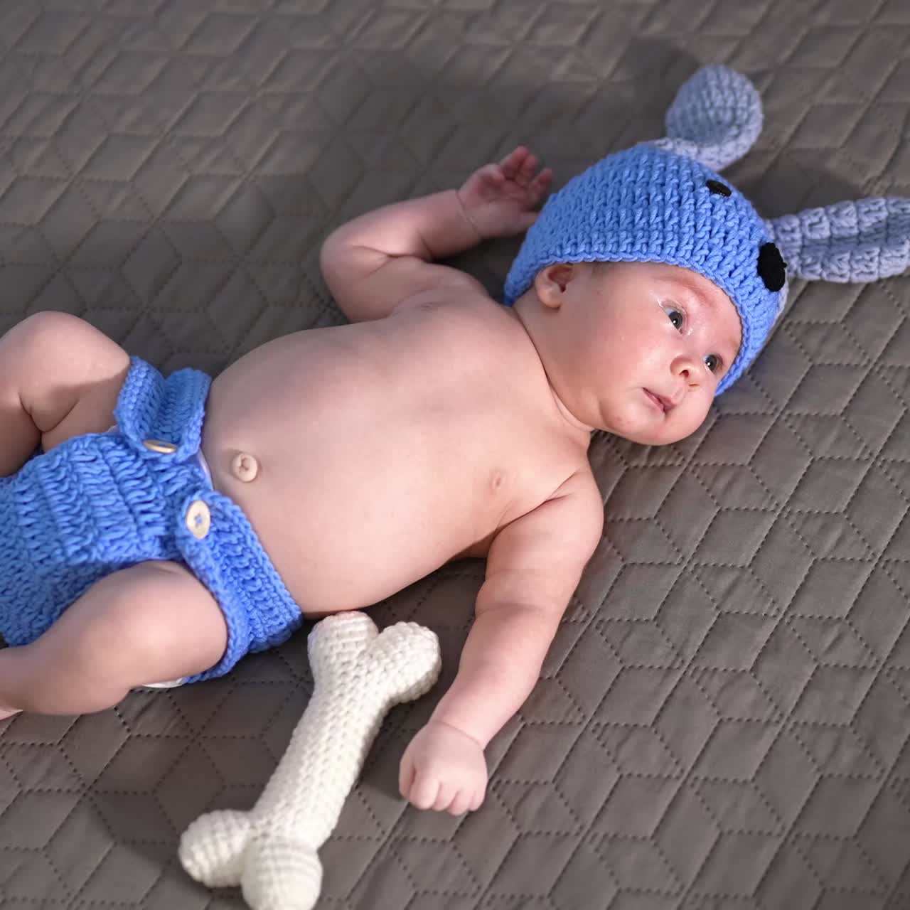 Cute baby in a funny blue suit lying on the bed. Puppy costume put on a newborn kid and funny soft knitted bone beside. View from above
