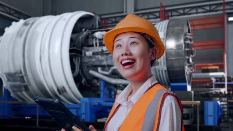close up side view of asian female engineer with safety helmet looking at the tablet in her hand and looking around while standing with airplane engine maintenance conducted