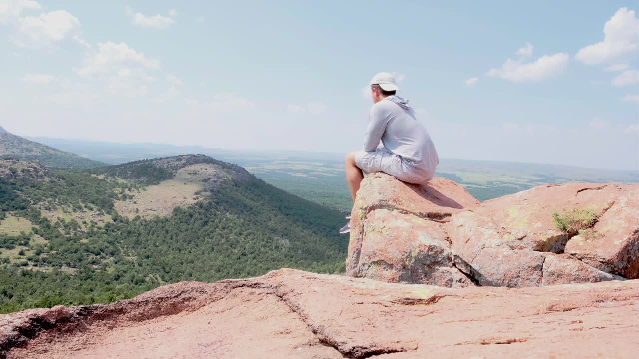 젊은 백인 남성 하이커가 오클라호마 주 마운트 스의 울창한 언덕을 내려다보는 바위 위에 앉아 있다.