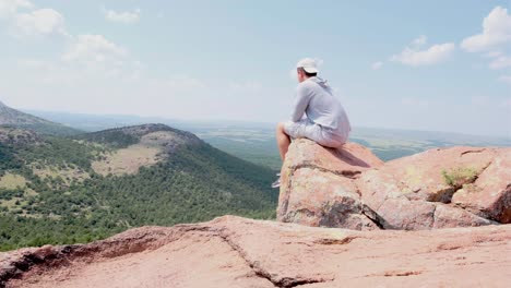 young, caucasian male hiker sitting down on a boulder overlooking lush hills at mount scott, oklahoma during daytime