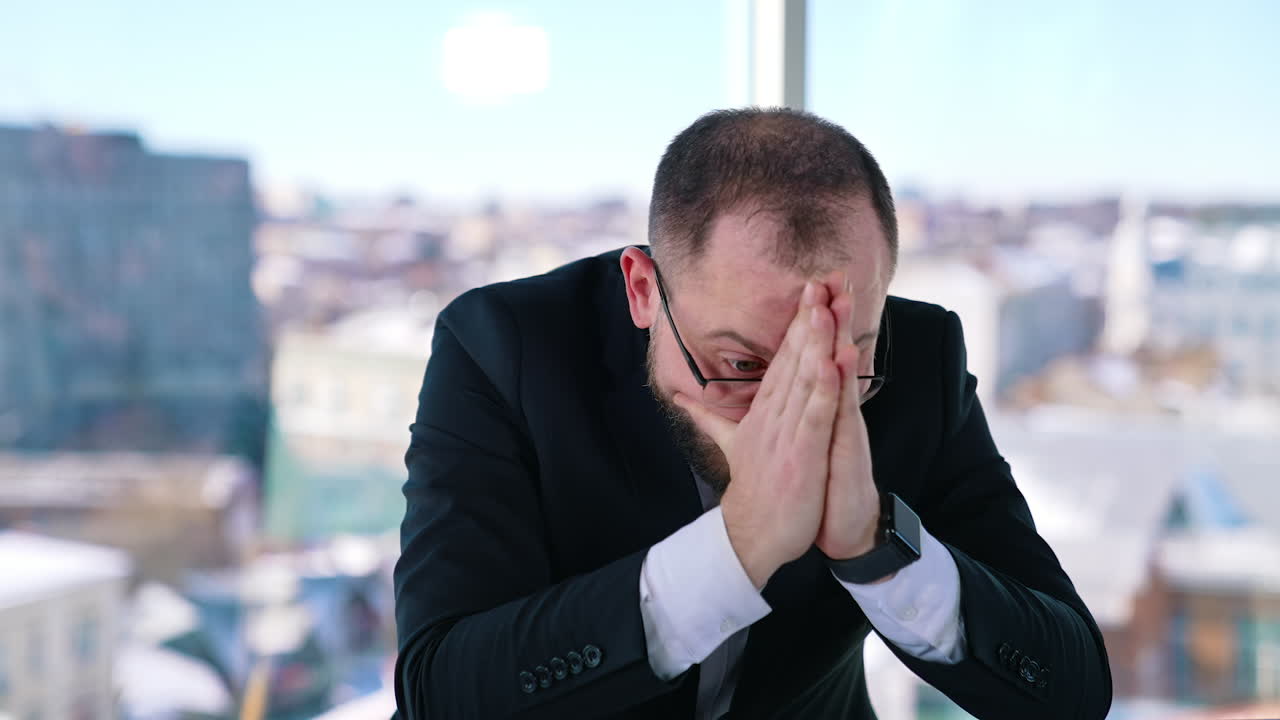 Depressed businessman sits at desk. Bearded middle aged man in glasses with his hands on head sitting at workplace and thinks. Portrait of sad businessman in office.