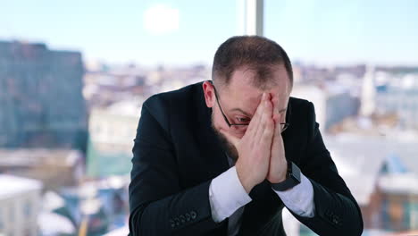 depressed businessman sits at desk. bearded middle aged man in glasses with his hands on head sitting at workplace and thinks. portrait of sad businessman in office.
