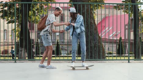 vista completa de una adolescente montando una patineta mientras su novio afroamericano la ayuda a mantener el equilibrio en la calle