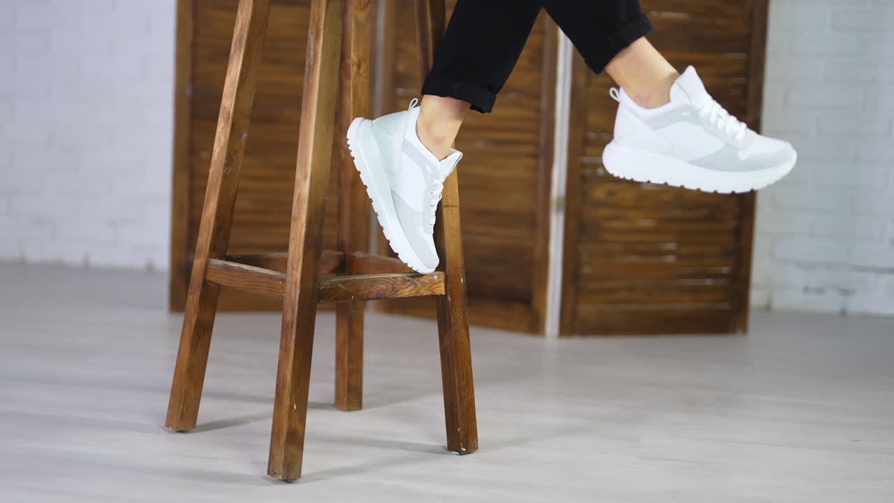 Model in black jeans comes to a wooden stool, sits on it and shows white new sneakers. Girl wave her legs to demonstrate fashionable footwear.