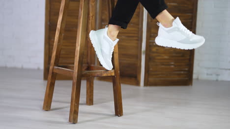 model in black jeans comes to a wooden stool, sits on it and shows white new sneakers. girl wave her legs to demonstrate fashionable footwear.