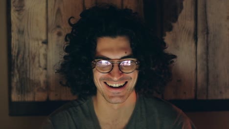 hombre caucásico de cabello rizado riendo durante una llamada de video iluminado por la pantalla de la computadora
