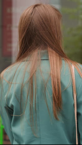 woman in elegant green jacket walks towards big glass door of shop building. young female person goes to buy products backside view slow motion