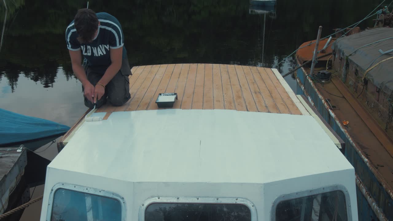 Young man paints half of wooden boat wheelhouse cabin with primer