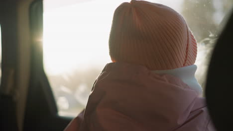 a little girl in a pink cap, jacket, and white winter scarf sits in a car, gazing thoughtfully through the window. the backlit scene creates a warm