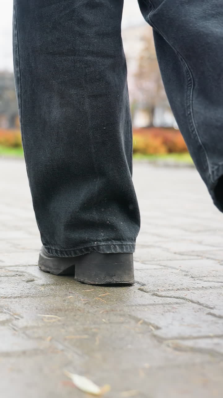 A human leg wearing black trousers and black boots walking slowly on outdoor paved path during calm overcast day, capturing foot movement and subtle step motion in quiet urban environment