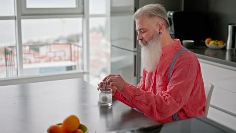 Side-view-of-an-elderly-man-with-gray-hair-and-a-lush-beard-in-a-pink-shirt-with-suspenders-pours-white-medicine-into-a-glass-of-water-and-stirs-it-with-a-spoon-in-a-modern-apartment