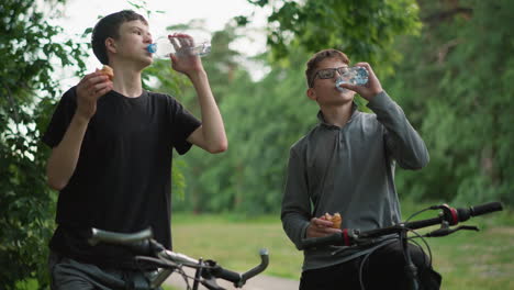 dos amigos en un paseo en bicicleta aplauden con sus botellas de agua antes de beber, sosteniendo bocadillos en sus manos, están rodeados de exuberante vegetación y árboles