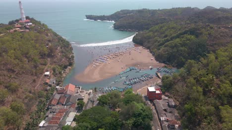Reveal-drone-shot-of-beach-with-harbor-that-full-of-fishing-boats,-white-lighthouse-on-the-top-of-cliff-and-sandy-area-to-playing---Baron-beach,-Indonesia