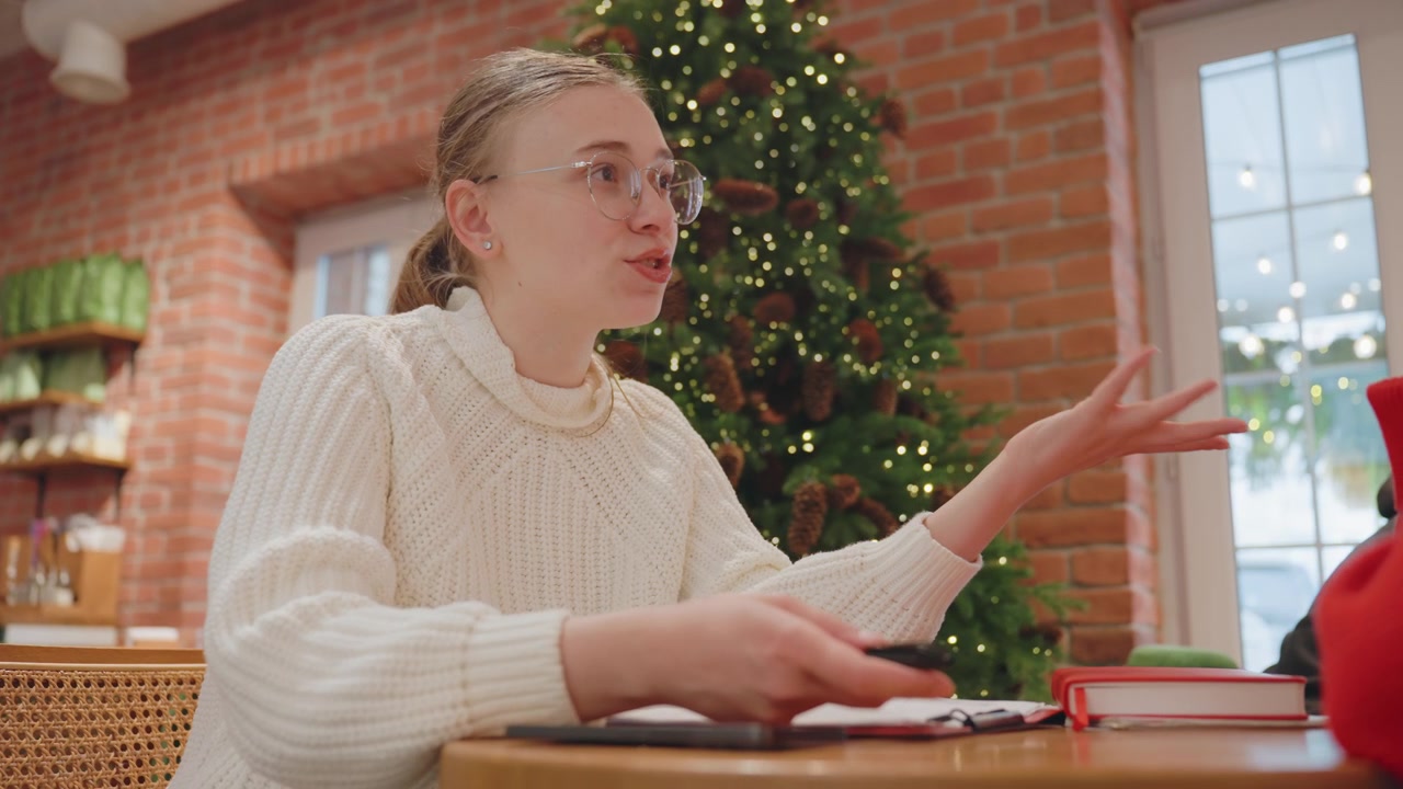 Nuori nainen silmälaseissa puhuu ja elehtii kahvilassa joulukuusen kanssa.