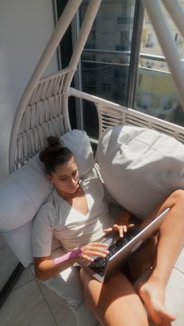 woman working remotely on a laptop in a hanging chair on a balcony