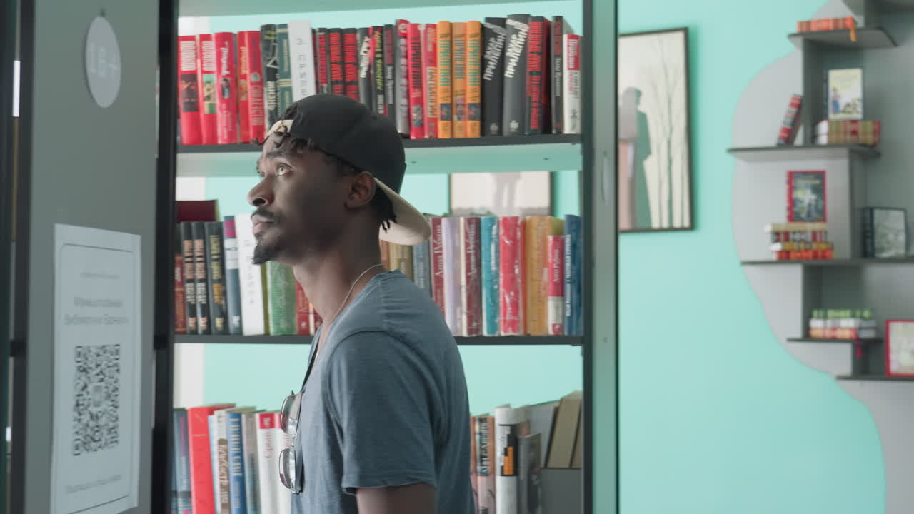 Side view of young man in casual t-shirt and backward cap exploring modern library, walking past tall bookshelves filled with colorful books