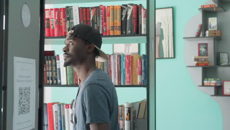 Side view of young man in casual t-shirt and backward cap exploring modern library, walking past tall bookshelves filled with colorful books